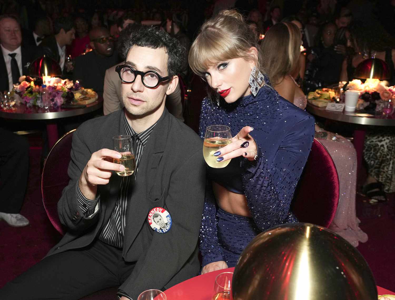 Jack antonoff wearing a black suit and Taylor Swift wearing blue out fit while holding glass